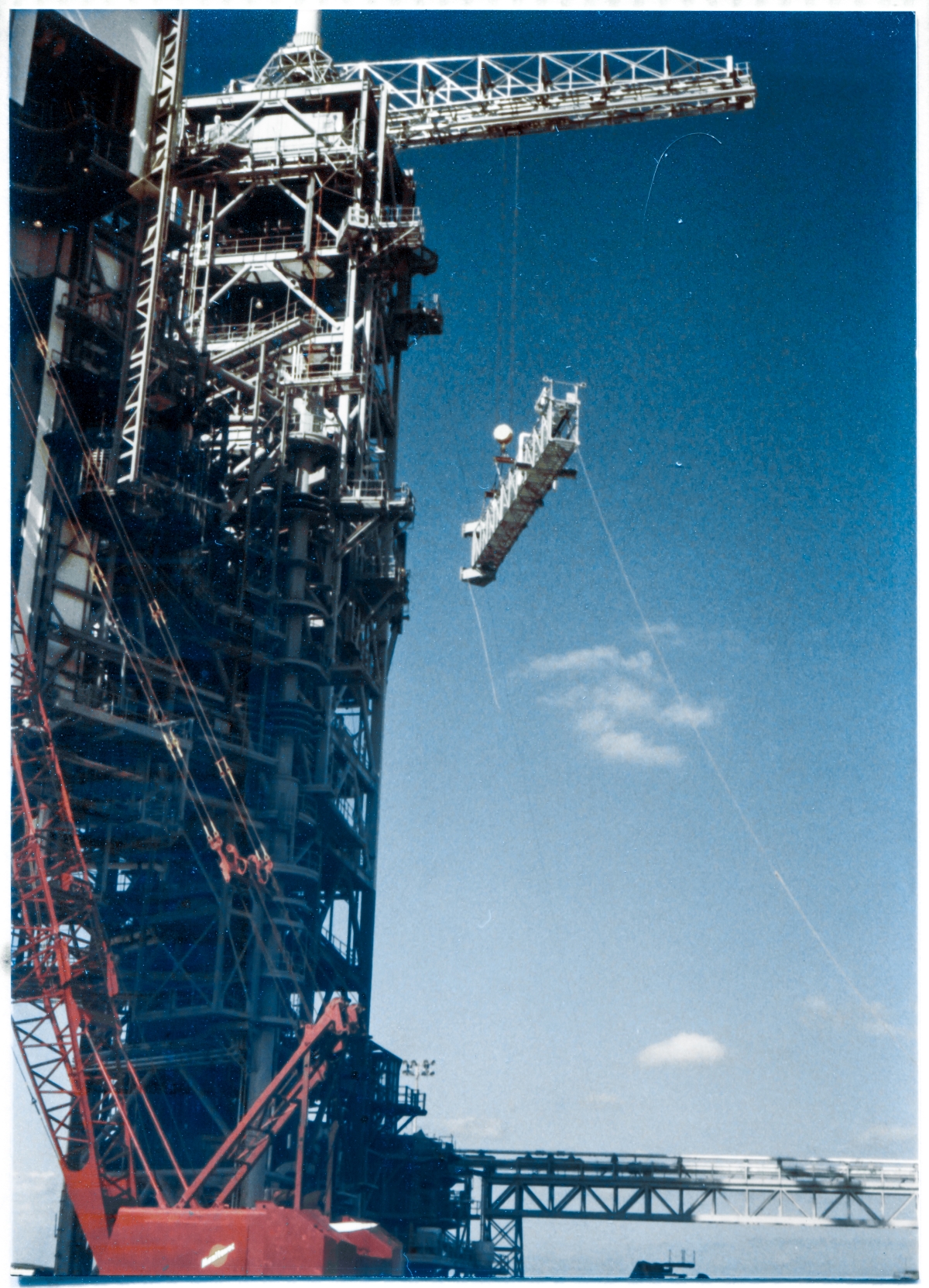 Image 107. Viewed from just south of the Flame Trench at Space Shuttle Launch Complex 39-B, Kennedy Space Center, Florida, the GOX Arm rises higher and higher into the sky, suspended beneath the Hammerhead Crane which is lifting it. Near the top of the Fixed Service Structure. From this distance, with the quality of the photograph that we've been given, it's impossible to tell with absolute certainty, but that is very likely Wade Ivey standing on the GOX Arm Lower Hinge Access Platform, white hardhat, dark jacket with just a bit of white across the shoulders, watching closely as the Arm approaches. Wade missed nothing, and as an ex-ironworker himself, he would go wherever the work was occurring, up on high steel, keeping a keen eye on things. This was a significant Lift, and he was in the thick of things the whole time. He never interfered. But he never went away, either. Photo by James MacLaren.